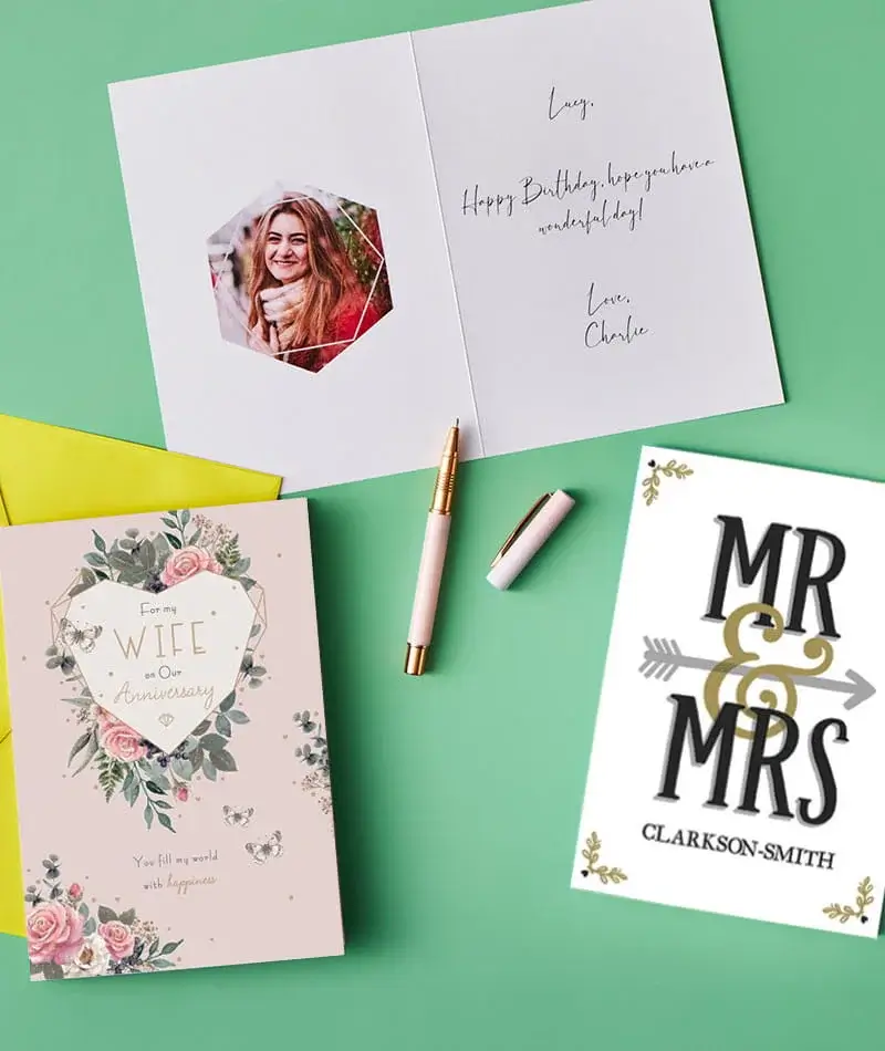 A selection of greeting cards displayed on a green surface, including an open birthday card with a handwritten message, a floral anniversary card, and a Mr and Mrs card, shown alongside pens and envelopes.